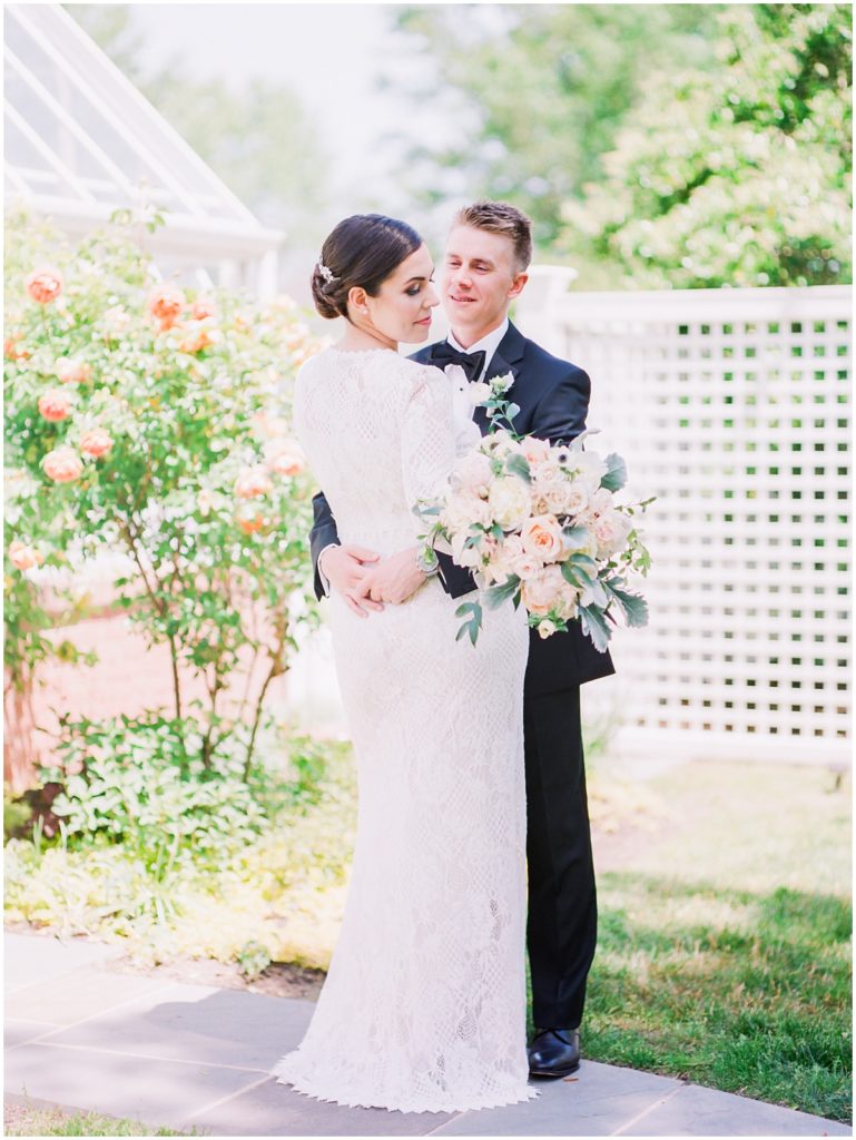 A brunette bride wearing a gorgeous full-sleeve, floor length lacey gown looks down at her flowers while her groom hugs her around the waist and smiles at her